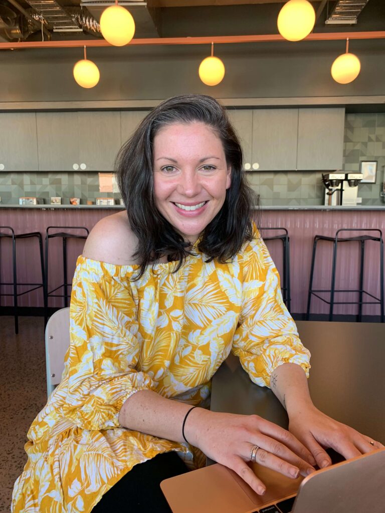 Clare in an off-the-shoulder yellow and white dress sitting at her laptop smiling at the camera
