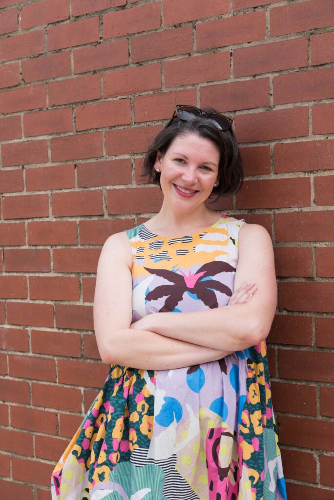 Clare standing against a brick wall with her arms folded wearing a colourful dress.