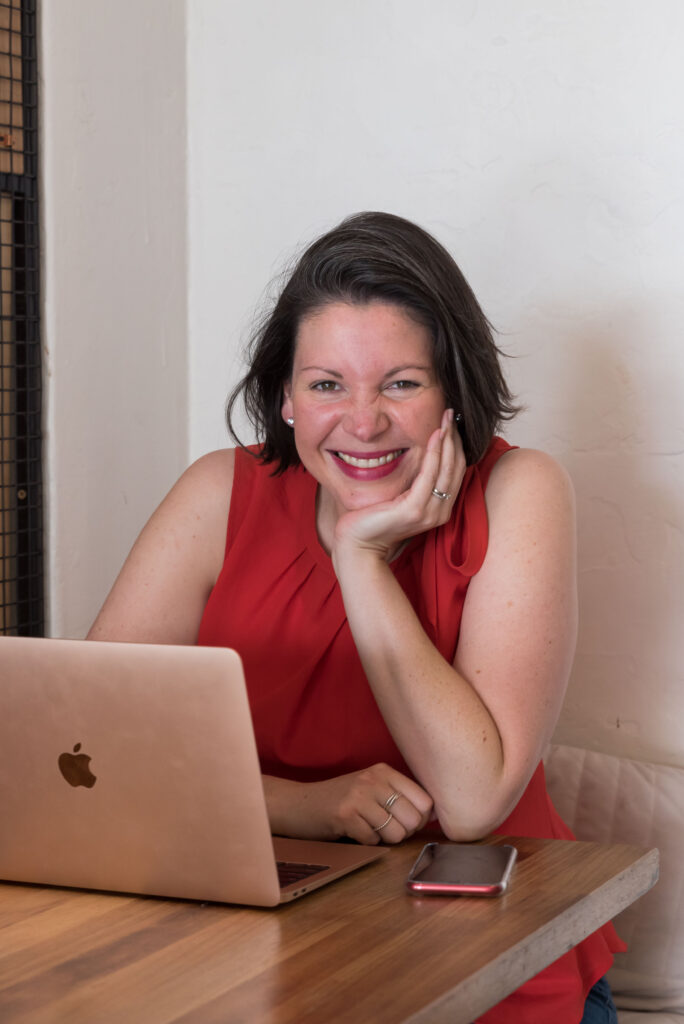 Clare sitting at a desk with her rose gold laptop resting her head in her hand and smiling