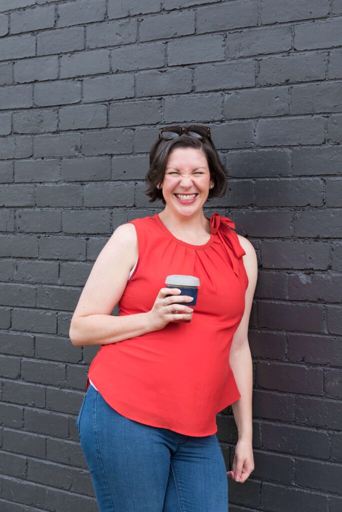 Clare wearing a red top standing against a black brick wall holding a coffee with a funny face