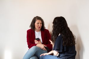Clare wearing a red jacket and jeans talking to someone who has their back turned to the camera.