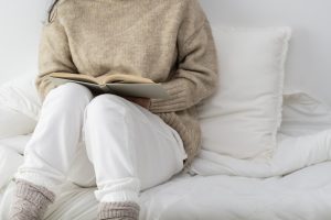 A woman wearing a cozy beige jumper and white pants sitting on a bed with white linen reading a book