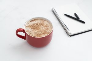 A cappucino in a red mug sitting next to a notepad and black pen