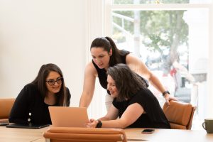 3 women wearing black in a light-filled studio staring at a laptop screen