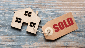 14:06A small cardboard cutout house on a weathered blue-grey wooden surface, with a kraft paper "SOLD" tag attached by twine. The Human Element logo and tagline appears in the bottom left corner.