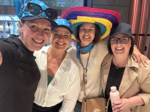 Four women smiling and laughing together in a selfie, wearing fun novelty hats — including blue bandanas, a vibrant rainbow-coloured sombrero, and a grey cap. They are dressed casually and appear to be at an indoor event, clearly having a great time together.